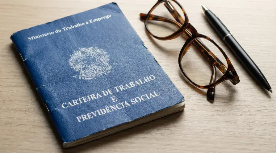 Carteira de Trabalho (CTPS) azul brasileira sobre uma mesa de madeira clara, ao lado de um par de óculos de grau e uma caneta preta