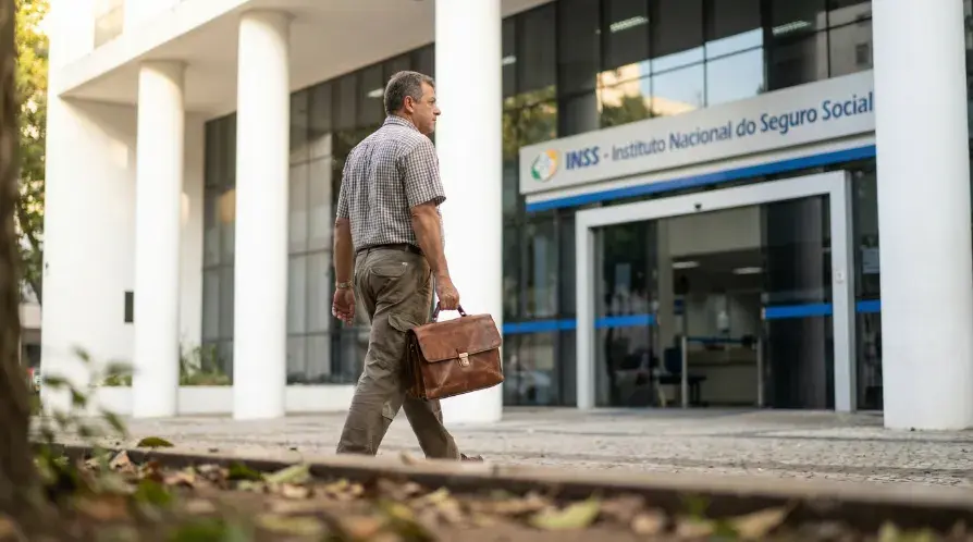 Foto de média distância de um homem de perfil, vestindo uma camisa xadrez e calça cáqui, carregando uma pasta de couro marrom enquanto caminha em uma calçada arborizada em direção à entrada de um prédio do INSS (Instituto Nacional do Seguro Social) com colunas brancas e fachada de vidro